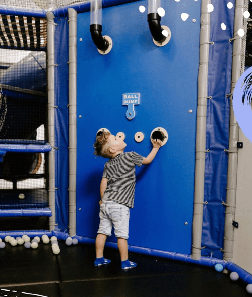 A child interacts with a blue ball dump wall in a play area, surrounded by soft play equipment and balls.