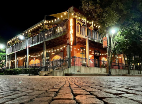 A brick building at night, adorned with string lights, featuring a balcony and surrounded by trees and cobblestone pavement.