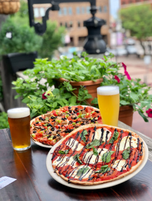Two pizzas on a wooden table, one topped with vegetables and the other with mozzarella and basil, alongside two glasses of beer.