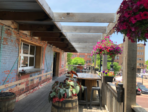 A rustic outdoor patio with wooden beams, colorful flowers, and a view of a vibrant street below.