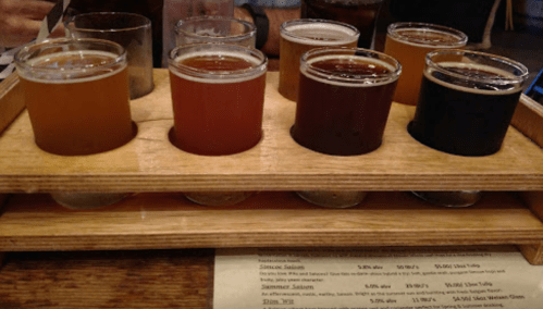 A wooden flight tray holds eight small glasses of different craft beers, showcasing various colors and styles.