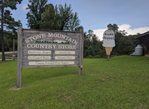 Sign for Stone Mountain Country Store, featuring offerings like sandwiches, ice cream, and local crafts, set in a green landscape.