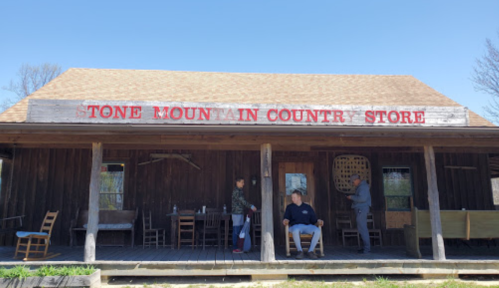 A rustic country store with a wooden facade, featuring chairs outside and people gathered on the porch.