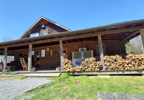 A rustic wooden cabin with a porch, stacked firewood, and a clear blue sky in the background.