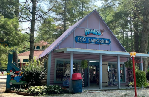 A colorful gift shop named "Rudi's Zoo Emporium" surrounded by trees, with outdoor displays and a red trash can.
