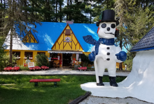 A large snowman statue stands in front of a colorful cottage with a blue roof, surrounded by trees and red wagons.