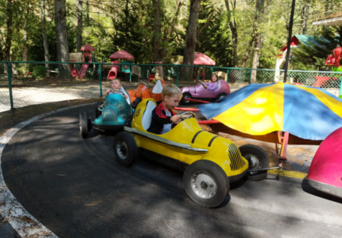 Two children ride colorful go-karts on a track surrounded by trees and playful structures.