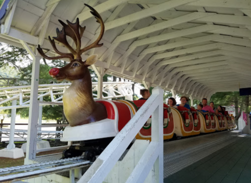 A roller coaster with a reindeer front car, passengers seated, under a white wooden structure.