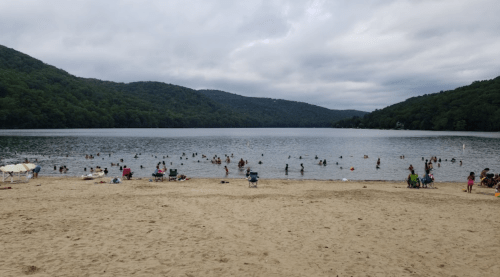 A sandy beach with people swimming in a lake, surrounded by green hills under a cloudy sky.