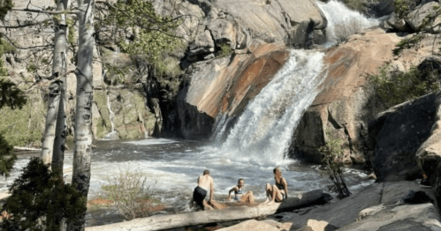 Three people sit on a log by a waterfall, surrounded by rocky terrain and greenery. Sunlight filters through the trees.