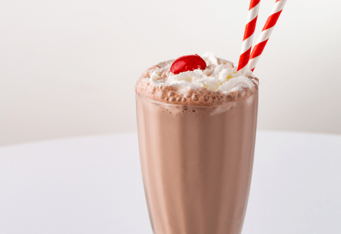 A tall glass of chocolate milkshake topped with whipped cream and a cherry, with red and white striped straws.