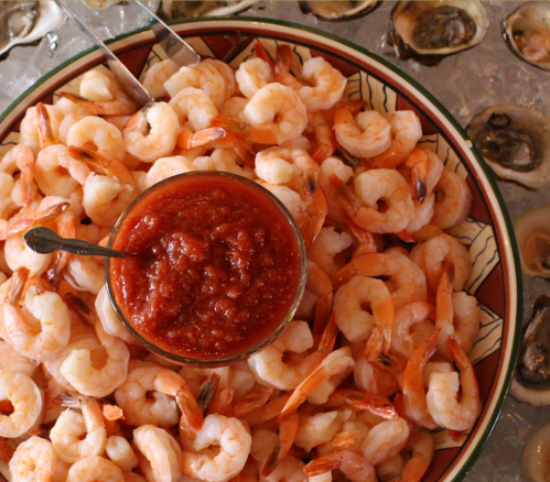 A platter of shrimp with a bowl of cocktail sauce in the center, surrounded by ice and oysters.