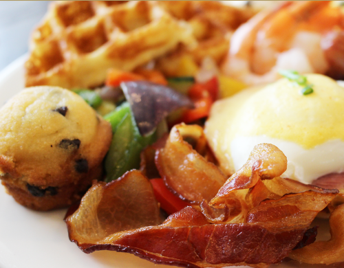A plate of breakfast featuring waffles, eggs Benedict, bacon, vegetables, and a blueberry muffin.