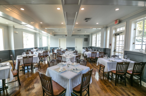 A spacious restaurant interior with neatly set tables, wooden chairs, and large windows letting in natural light.