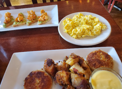 A table with three plates: shrimp on toast, scrambled eggs, and a dish of potatoes with two crispy cakes and sauce.
