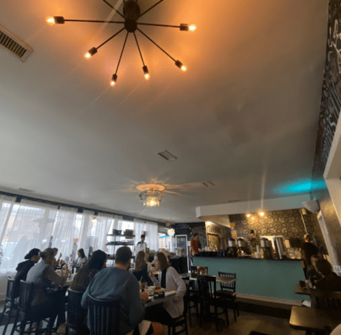 A cozy restaurant interior with diners at tables, a modern chandelier, and a bar area in the background.