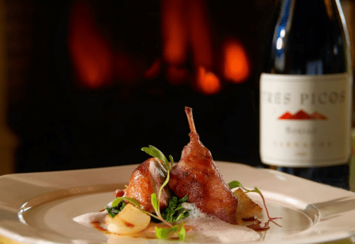 A gourmet dish featuring a crispy piece of meat garnished with greens, served with a bottle of wine in the background.