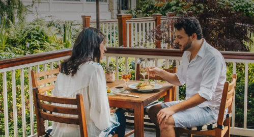 A couple enjoys a meal together on a patio, surrounded by greenery, with drinks and food on the table.