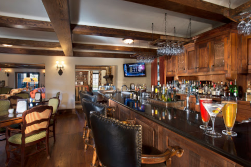 A cozy bar area with wooden beams, a polished counter, and colorful cocktails on display. Comfortable seating nearby.