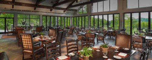 A spacious restaurant with large windows, wooden beams, and elegantly set tables overlooking a green landscape.
