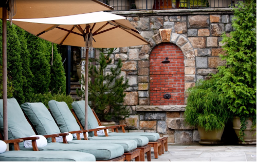 Lounge chairs with green cushions under umbrellas, beside a stone wall with a brick arch and greenery.