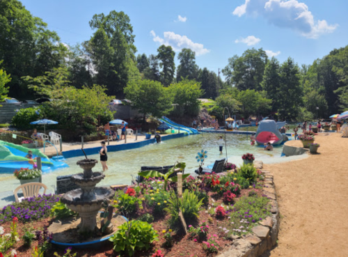 A sunny water park scene with slides, a pool, and colorful flowers, filled with people enjoying the day.
