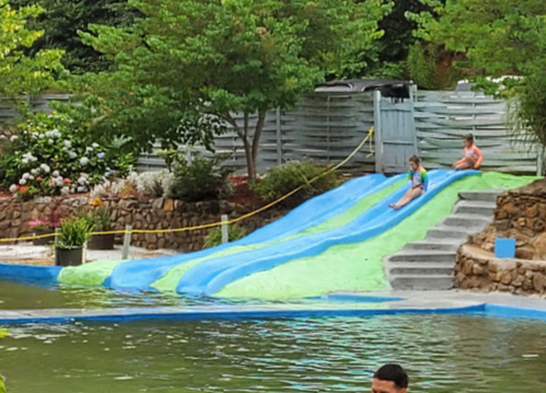 Two children slide down a colorful water slide into a pool, surrounded by greenery and a stone wall.