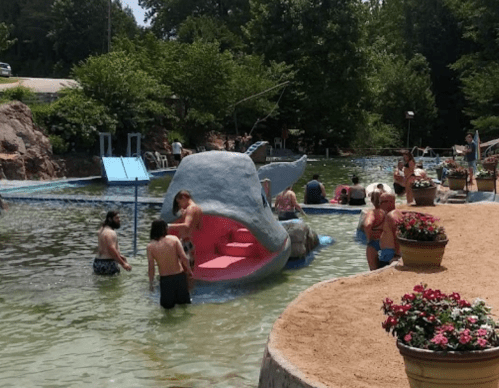 A whimsical whale sculpture in a swimming area, with people enjoying the water and greenery around.