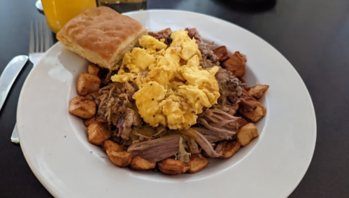 A plate of pulled pork topped with scrambled eggs, served with crispy potatoes and a biscuit on the side.