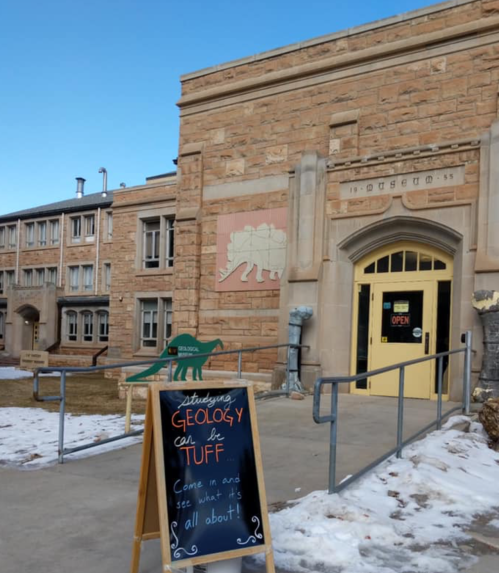 A stone building with a dinosaur mural and a sign inviting visitors to learn about geology. Snow on the ground.