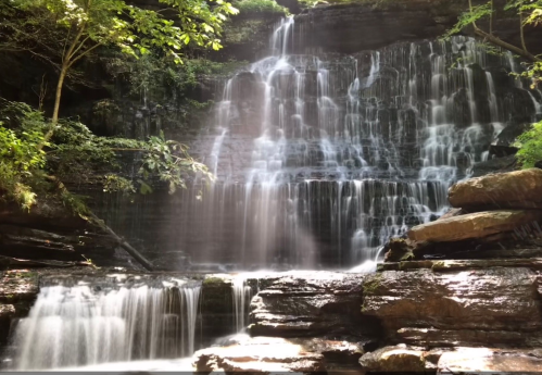 A serene waterfall cascading over rocks, surrounded by lush green trees and soft mist in a tranquil forest setting.
