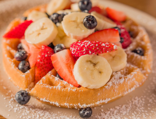 A waffle topped with sliced bananas, strawberries, and blueberries, dusted with powdered sugar.