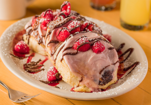 A plate of sweet rolls topped with raspberry icing, fresh raspberries, chocolate drizzle, and powdered sugar.