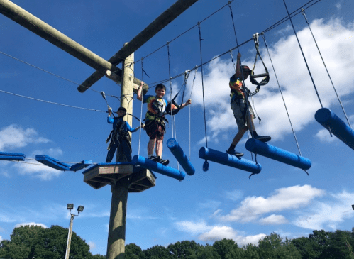 Two children navigate a rope course with blue obstacles, suspended high above the ground, under a clear blue sky.