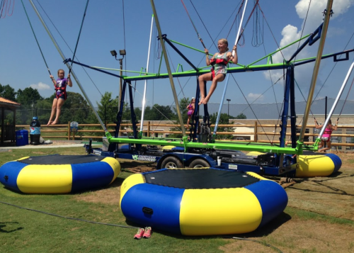 Children enjoying a bungee trampoline setup, bouncing on colorful inflatable pads under a clear blue sky.