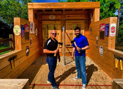 Two men stand in an outdoor axe-throwing venue, each holding an axe, smiling at the camera. Wooden walls are decorated with signs.