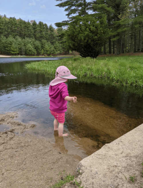 A child in a pink outfit and hat stands in shallow water by a sandy shore, surrounded by greenery and trees.
