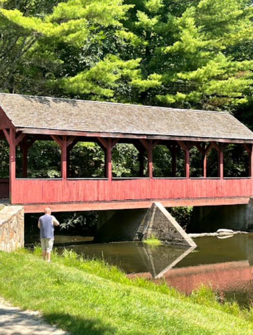 A red covered bridge spans a calm river, with a person standing nearby on the grassy bank. Trees line the background.