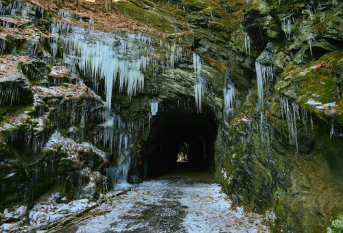 A dark tunnel in a rocky landscape, with icicles hanging from the edges and snow on the ground.