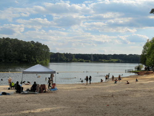 A sandy beach by a lake with people swimming, relaxing, and a tent set up under a partly cloudy sky.