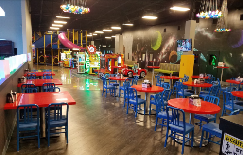 Brightly colored indoor play area with tables, arcade games, and a playground structure in a spacious setting.