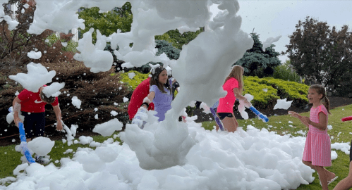 A group of children play in a large pile of foam, laughing and having fun in a garden setting.