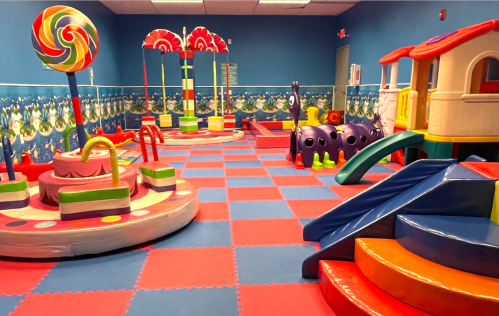 Colorful indoor playroom with a slide, climbing structures, and a carousel, featuring bright mats and playful decor.