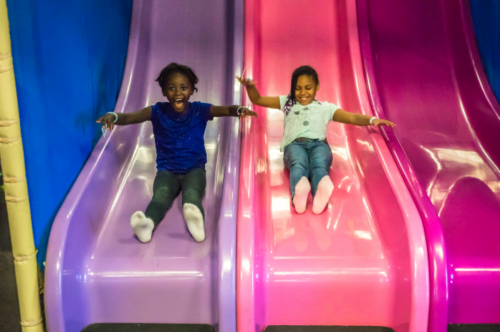 Two children joyfully sliding down colorful slides in a play area, one in blue and the other in pink.