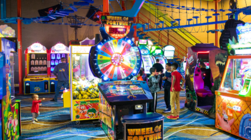 A colorful arcade filled with games, featuring children playing and a "Wheel of Fortune" game in the foreground.