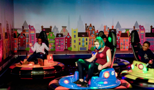Children enjoying bumper cars in a colorful indoor amusement setting with vibrant cityscape backdrop.