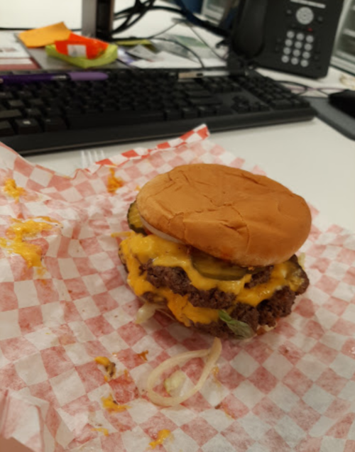 A messy cheeseburger with two patties, melted cheese, pickles, and onions on a checkered paper, set on a desk.