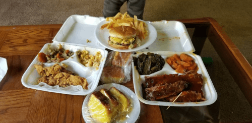 A takeout meal featuring fried chicken, ribs, a burger, fries, and various sides on a wooden table.