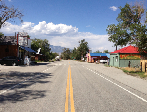 A quiet rural road lined with small shops and trees under a blue sky with scattered clouds.