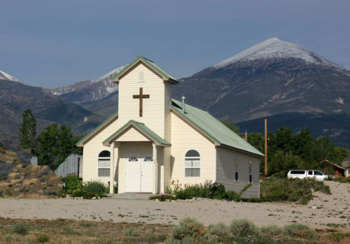 A small church with a cross on its facade, set against a backdrop of mountains and clear blue sky.